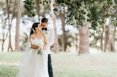 a bride and groom are standing in the grass under some trees with their arms around each other