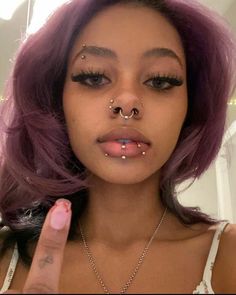 a close up of a person with purple hair and piercings on her nose pointing to the camera