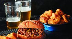 a sloppy joe sandwich on a plate next to some fried potatoes and beer in glasses