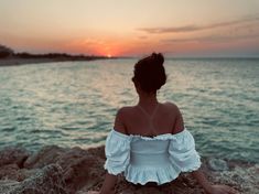 a woman sitting on the rocks looking out at the water and setting sun in the distance