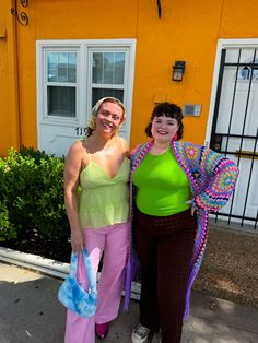 two women standing next to each other in front of a building with orange walls and white doors