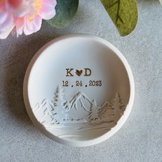 a white bowl with the words k d on it sitting next to flowers and leaves