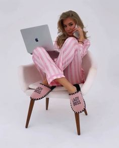 a woman in pink and white pajamas sitting on a chair using a laptop computer with her legs crossed