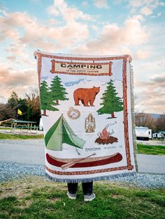 a person standing in the grass holding up a blanket with images of camping and trees on it