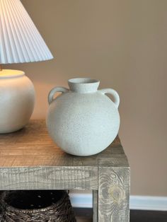 a white vase sitting on top of a wooden table next to a lamp and wicker basket