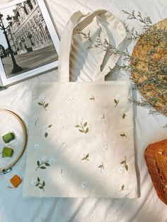 a white bag sitting on top of a bed next to a basket with flowers in it