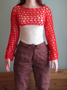 a woman standing in front of a bed wearing brown pants and a white top with red crochet on it