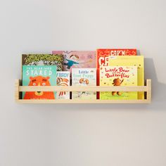 A natural wood wall shelf filled with children's books, mounted on a light gray wall. The shelf holds colorful book covers, making it an inviting and organized space for young readers. Soft Toy Storage, Floating Bookshelf, Nursery Bookshelf, Kids Shelves, Nursery Trends, Wall Mounted Bookshelves, Nursery Shelves, Prop Stylist, Wall Bookshelves