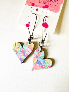 two heart shaped paper earrings hanging from hooks on a white surface next to a card