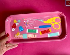 a person holding a pink tray with sewing supplies on it and scissors in the middle