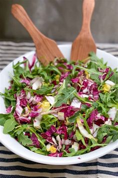 a salad in a white bowl with wooden spoons on the side and a striped tablecloth