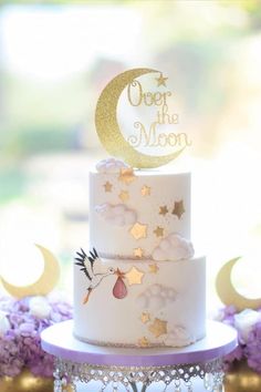 a three tiered white cake with gold stars and moon on top, in front of purple flowers