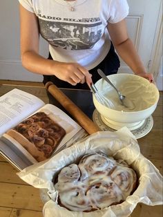 a woman is making cinnamon rolls in the kitchen with a book on the table next to her