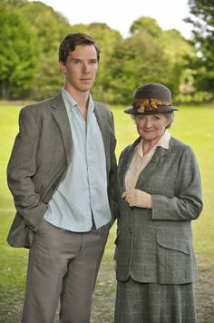 two people standing next to each other in front of trees and grass, one wearing a hat