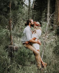 a man and woman kissing in the woods with pine trees behind them, surrounded by tall grass