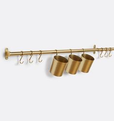 three brass cups hanging from a metal bar with hooks on the side and two empty mugs in front