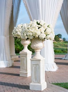 two white urns with flowers in them on a brick walkway next to drapes