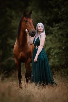 a woman in a long green dress standing next to a brown horse with her hand on it's face