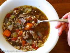 a person holding a spoon in a bowl filled with meat and barley soup on top of a wooden table