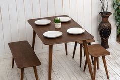 a wooden table with four white plates and two brown chairs next to a vase on the floor