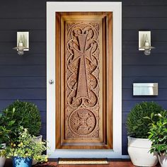 an ornate wooden door with potted plants on the front porch and blue house behind it