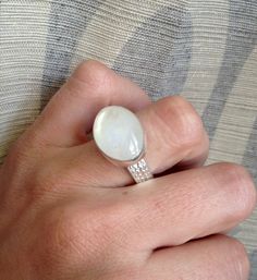 An elegant crystal white large oval Rainbow Moonstone has been set in sterling silver. The ring band is handmade from a fancy patterned sterling silver thick wire. This moonstone is pearly white and shines on its own with hues of pink, blue, and even orange. The luminescent quality of the moonstone makes this ring beautifully unique and elegant. The stone is 13mm x 18mm in size with a height of about 8mm. This moonstone can be set vertically (North to South) rather than horizontally as shown. Ju Modern Oval Moonstone Ring With Large Stone, Modern Handmade Oval Moonstone Ring, Handmade Oval Moonstone Ring In Modern Style, Handmade Modern Oval Moonstone Ring, Unique White Oval Cabochon Rings, Modern Oval Moonstone Jewelry, White Oval Cabochon Jewelry With Large Stone, White Oval Moonstone Ring With Large Stone, White Oval Ring With Large Stone