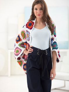 a woman standing in front of a white wall wearing black pants and a colorful crocheted jacket