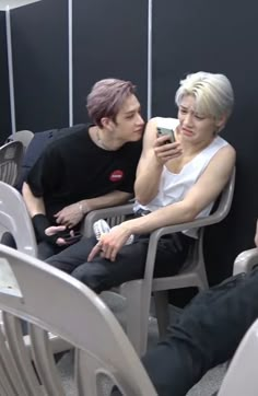 two people are sitting in chairs and one is looking at his cell phone while the other looks on