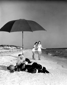 an old black and white photo of two women on the beach with an umbrella over them