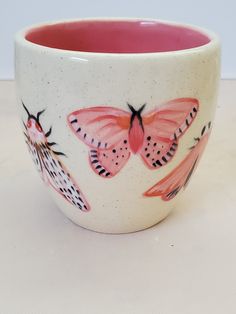 a pink and white cup with butterflies painted on it's sides, sitting on a table