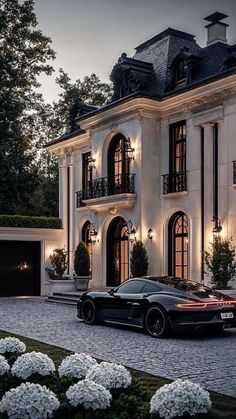 two black sports cars parked in front of a large white house at night with lights on