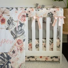 a white crib with pink and green floral bedding on it, next to a potted plant