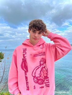 a young man standing next to the ocean wearing a pink hoodie with an anime character on it