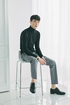 a man sitting on top of a white chair in a black turtle neck sweater and slacks