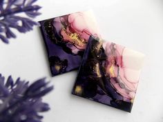 two square coasters sitting on top of a white table next to purple and gold flowers