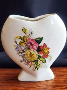 a white heart shaped vase with flowers painted on the front and sides, sitting on a wooden table