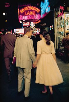 a man and woman holding hands walking down the street in front of a neon sign