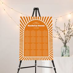 an orange and white wedding seating chart on a easel with lights in the background