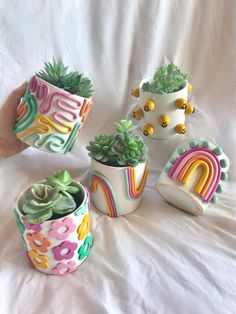 four ceramic planters with plants in them on a white sheeted surface, one has a rainbow design and the other has a potted cactus