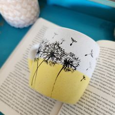 a coffee cup with dandelions on it sitting next to an open book