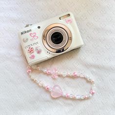 a white camera sitting on top of a bed next to a pink beaded necklace