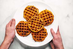 three waffles on a white plate being held by two hands over a marble table