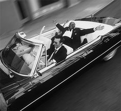 a man in a tuxedo is waving from the back of a convertible car