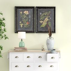 two framed pictures hang above a dresser with drawers and vases on it's sides