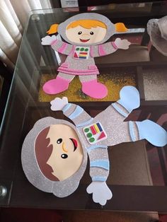 two paper dolls on top of a glass table in the shape of an astronaut's suit