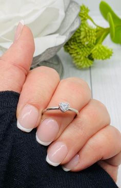 a woman's hand holding a ring with a heart shaped diamond on it and flowers in the background