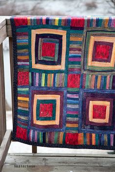 a multicolored quilt is hanging on a wooden bench in front of snow covered trees