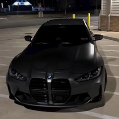 the front end of a black car parked in a parking lot at night with its lights on