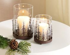 two candles sitting on top of a white table next to pine cones and evergreen branches