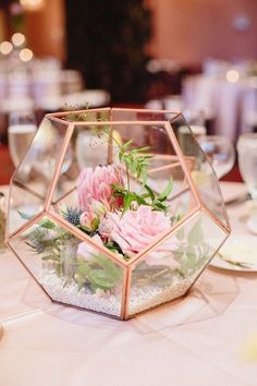 a glass vase filled with pink flowers and greenery on top of a white table cloth